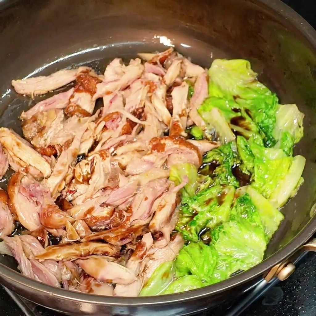 Zerkleinertes Hähnchen und Salat werden mit dunkler Sojasauce in der Pfanne vermischt.