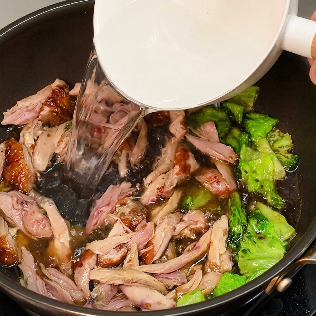 Heißes Wasser wird aus einer weißen Schüssel in die Pfanne mit gewürztem Hähnchen und Salat gegossen, um eine Brühe zu bilden.