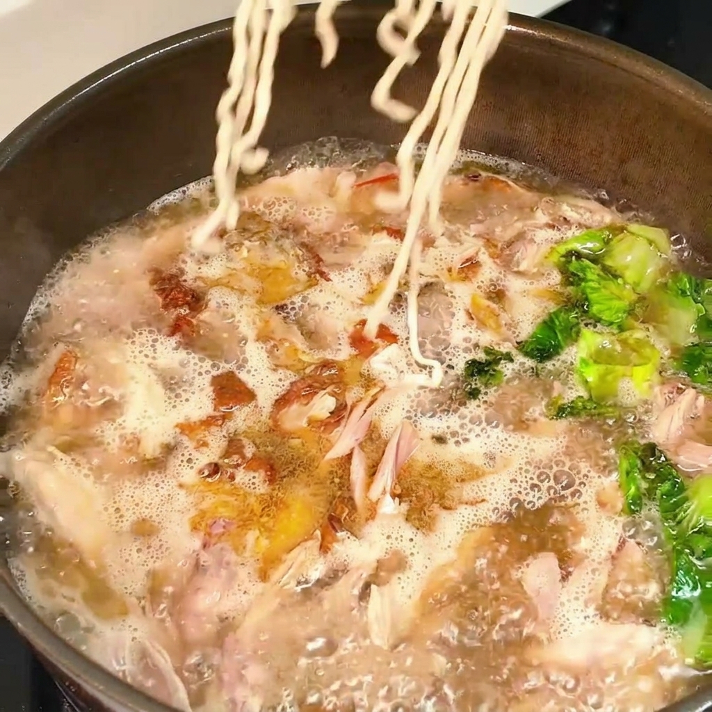 Ein Block trockene Wellennudeln wird in eine Pfanne mit sprudelnder Hähnchen-Salat-Brühe gegeben.