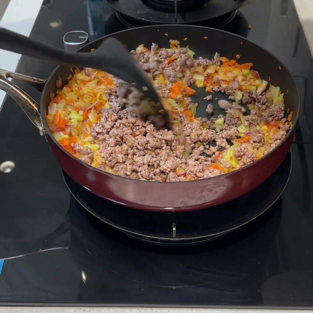 Gebratenes Hackfleisch und gewürfeltes Gemüse werden mit einem Pfannenwender in einer Pfanne vermengt.