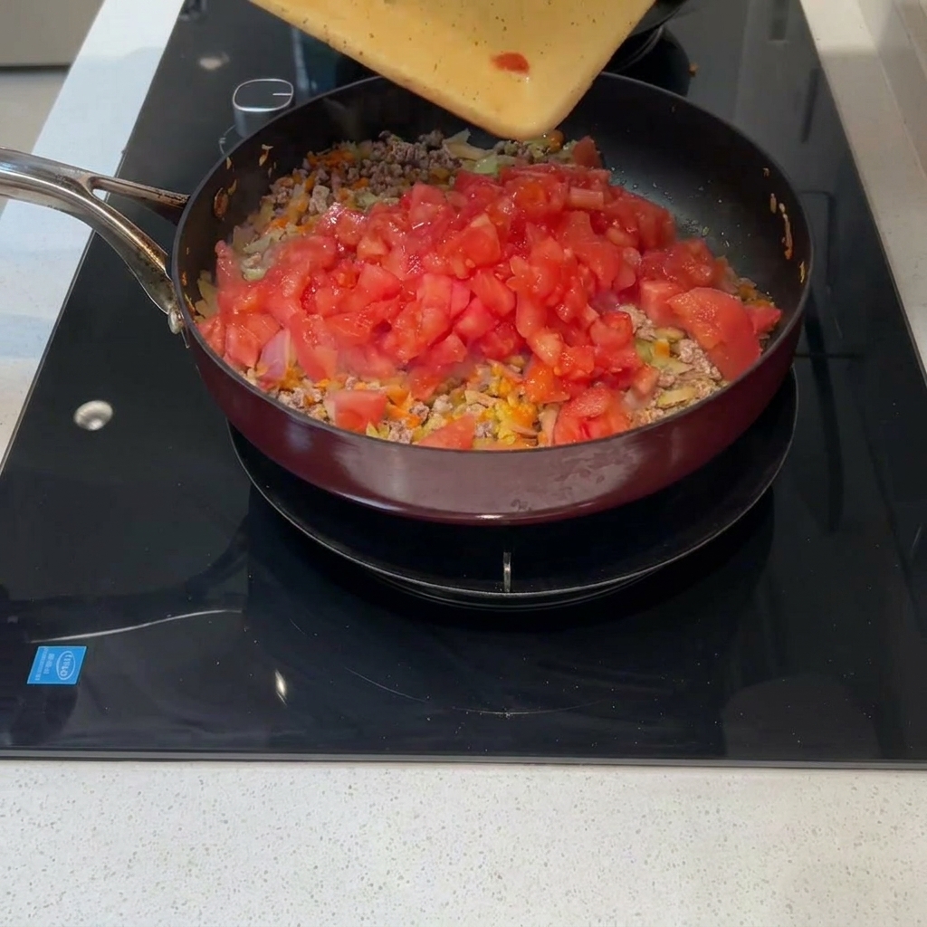 Frisch gewürfelte Tomaten werden von einem Schneidebrett in die Pfanne mit Fleisch und Gemüse gegeben.