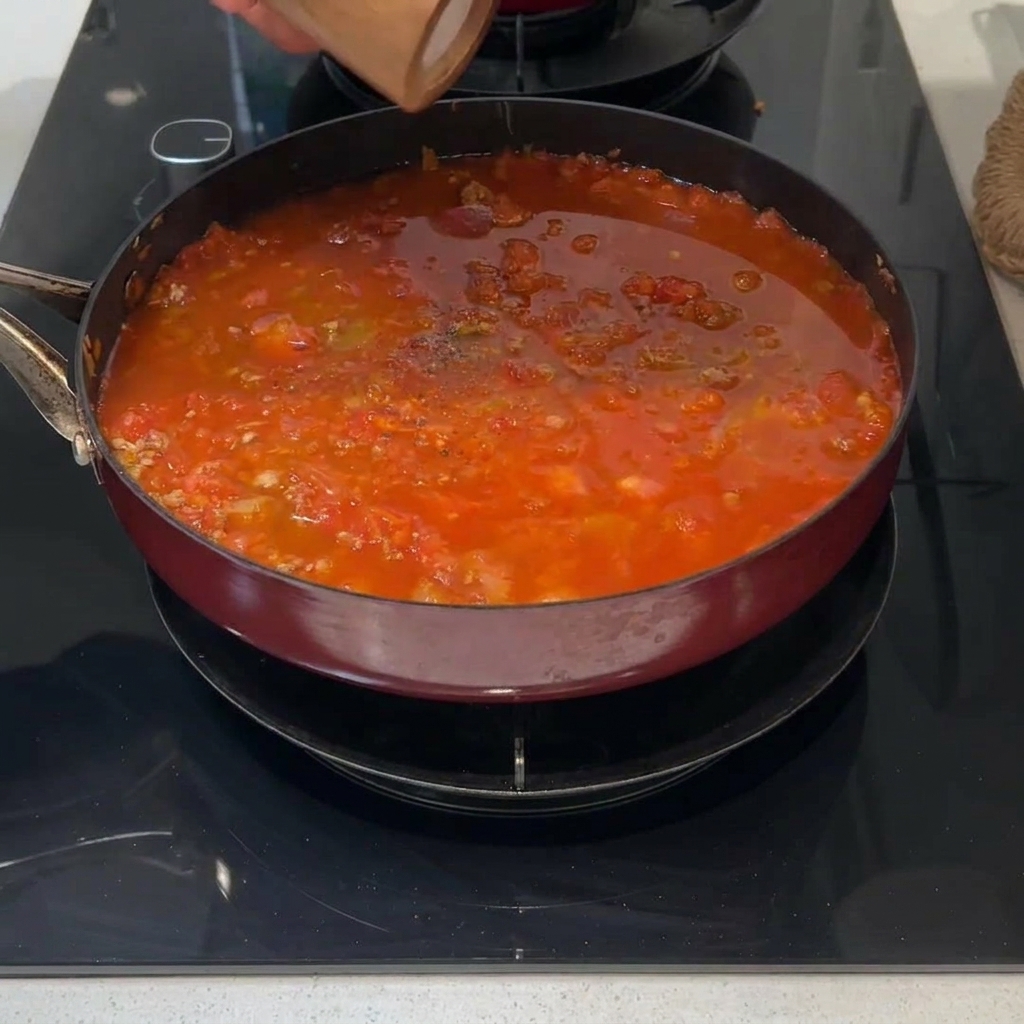 Ein frischer grüner Rosmarinzweig liegt auf der Oberfläche einer köchelnden roten Fleischsauce.