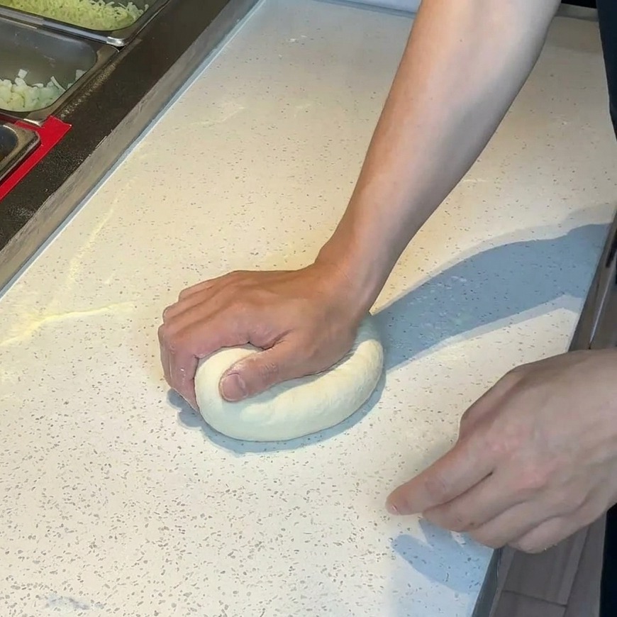 Hände drücken eine runde Kugel Pizzateig auf einer bemehlten weißen Arbeitsfläche flach.
