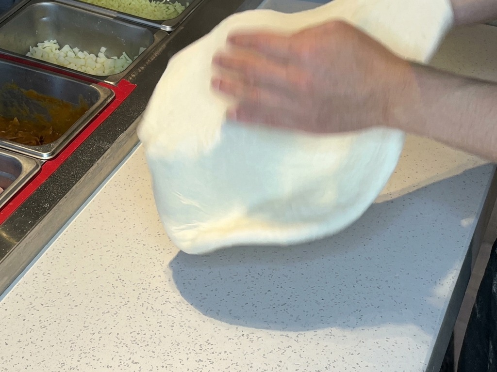 Zwei Hände heben und werfen gedehnten Pizzateig in die Luft, um überschüssiges Mehl zu entfernen.