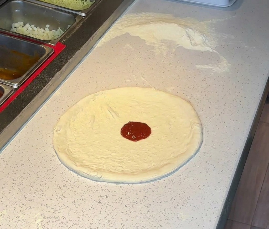 Ein Klecks reichhaltiger roter Tomatensoße in der Mitte eines gedehnten Pizzabodens auf einer Arbeitsplatte.
