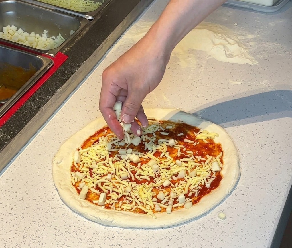 Eine Hand streut geriebenen weißen Käse über einen mit Tomatensoße bestrichenen Pizzaboden.