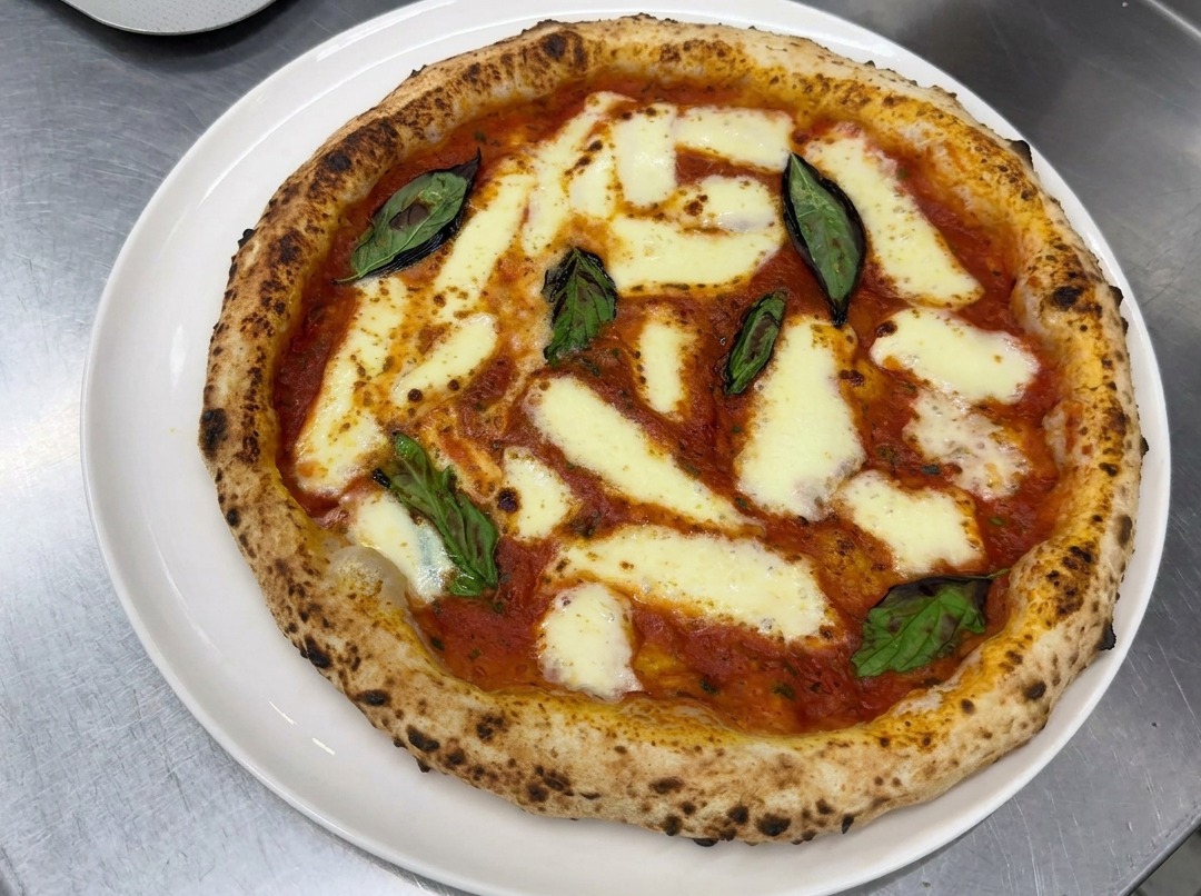 Hände dehnen vorsichtig eine Kugel Pizzateig auf einer bemehlten Arbeitsfläche zu einem runden Boden.