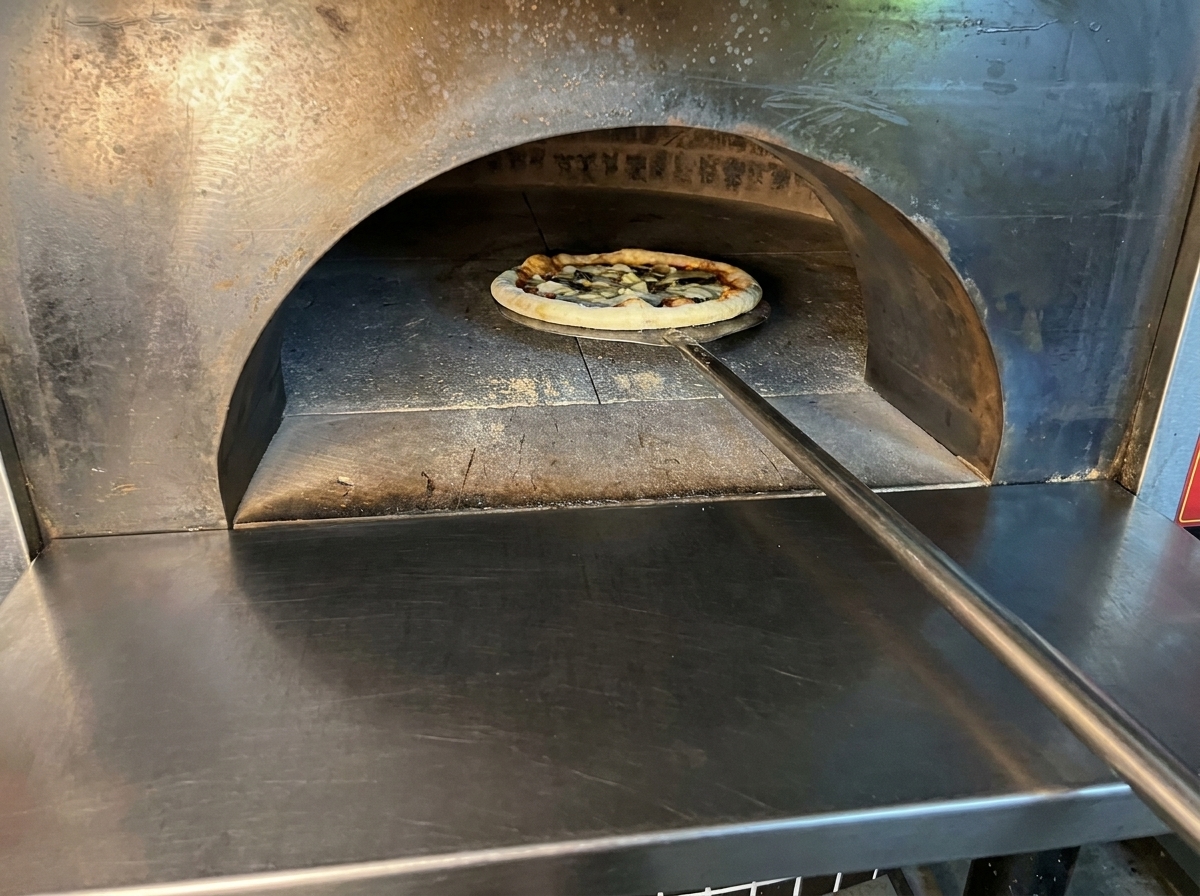 Zusammengebaute rohe Pizza, die mit einem langen Metallschieber in einen heißen Holzofen geschoben wird.
