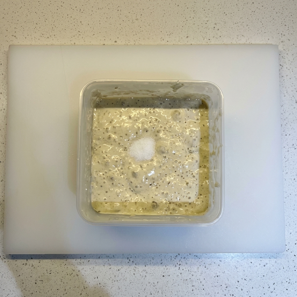 Salz und ein Häufchen Mehl liegen auf einer blubbernden, fermentierten Teigmischung in einem quadratischen Behälter.