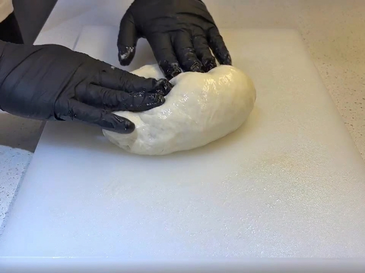 Hände mit schwarzen Handschuhen falten eine weiche, geölte Teigkugel auf einem weißen Schneidebrett.
