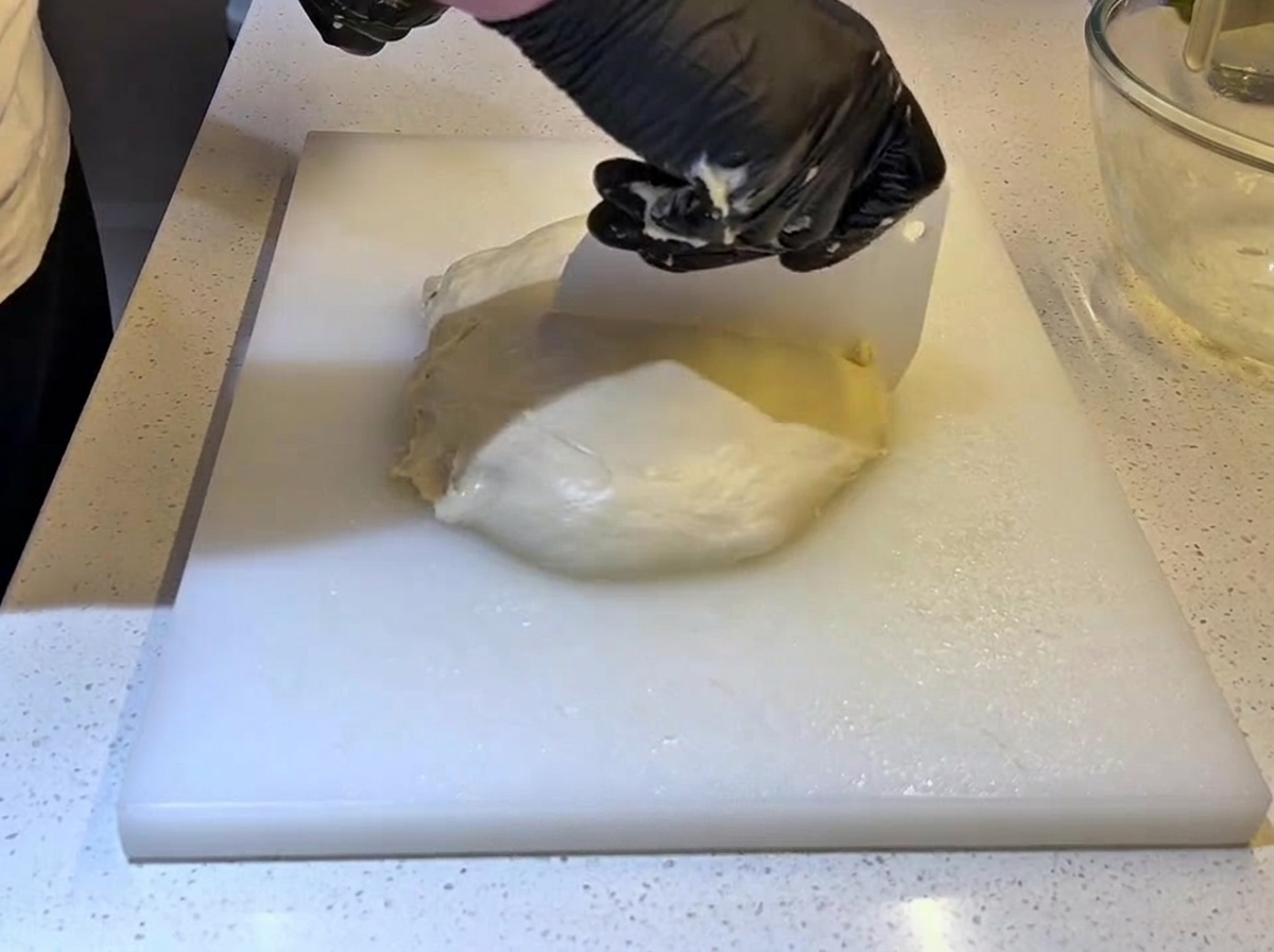 Hände in schwarzen Handschuhen verwenden eine weiße Teigkarte aus Plastik, um ein großes Stück Teig in zwei Hälften zu schneiden.
