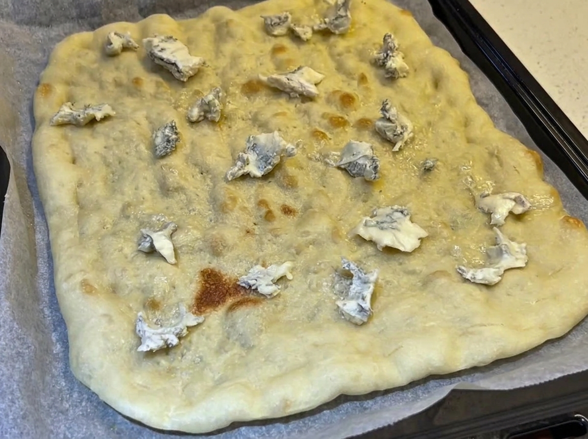 Ein vorgebackener, rechteckiger Fladenbrotboden, der mit Blauschimmelkäse-Bröckeln belegt ist, auf einem mit Backpapier ausgelegten Backblech.