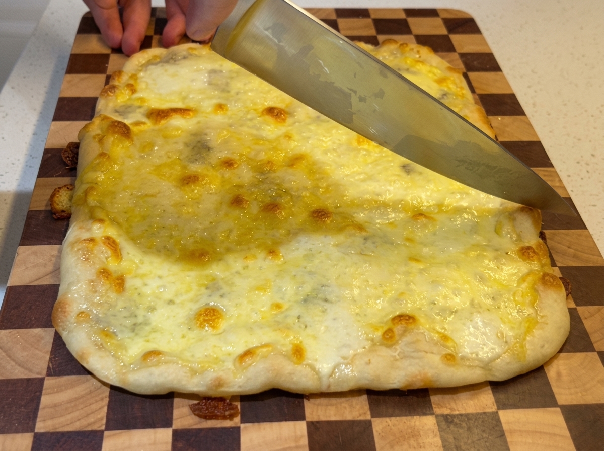 Ein großes Kochmesser schneidet durch eine frisch gebackene, blubbernde rechteckige Vier-Käse-Pizza auf einem karierten Holzschneidebrett.