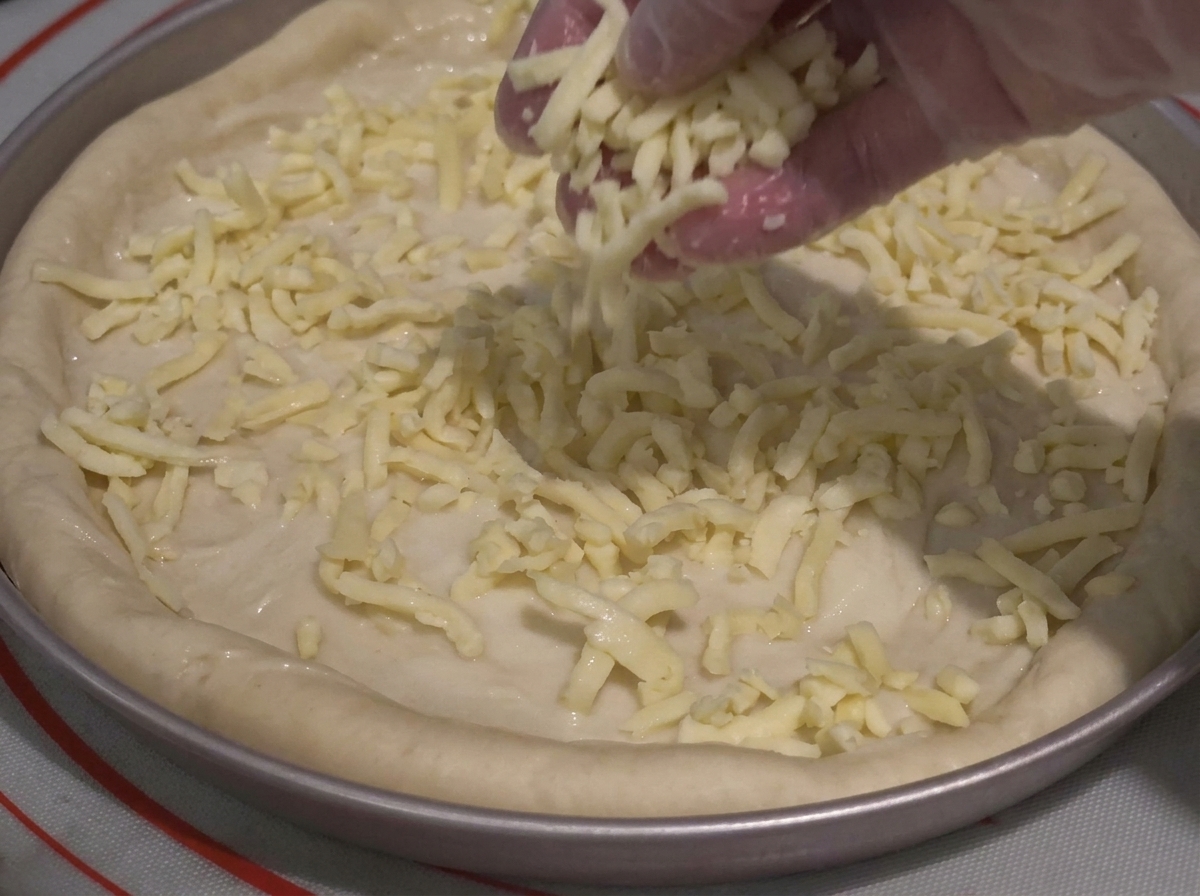 Eine Hand bestreut den mit Sauce bedeckten Teig großzügig mit geriebenem Käse.