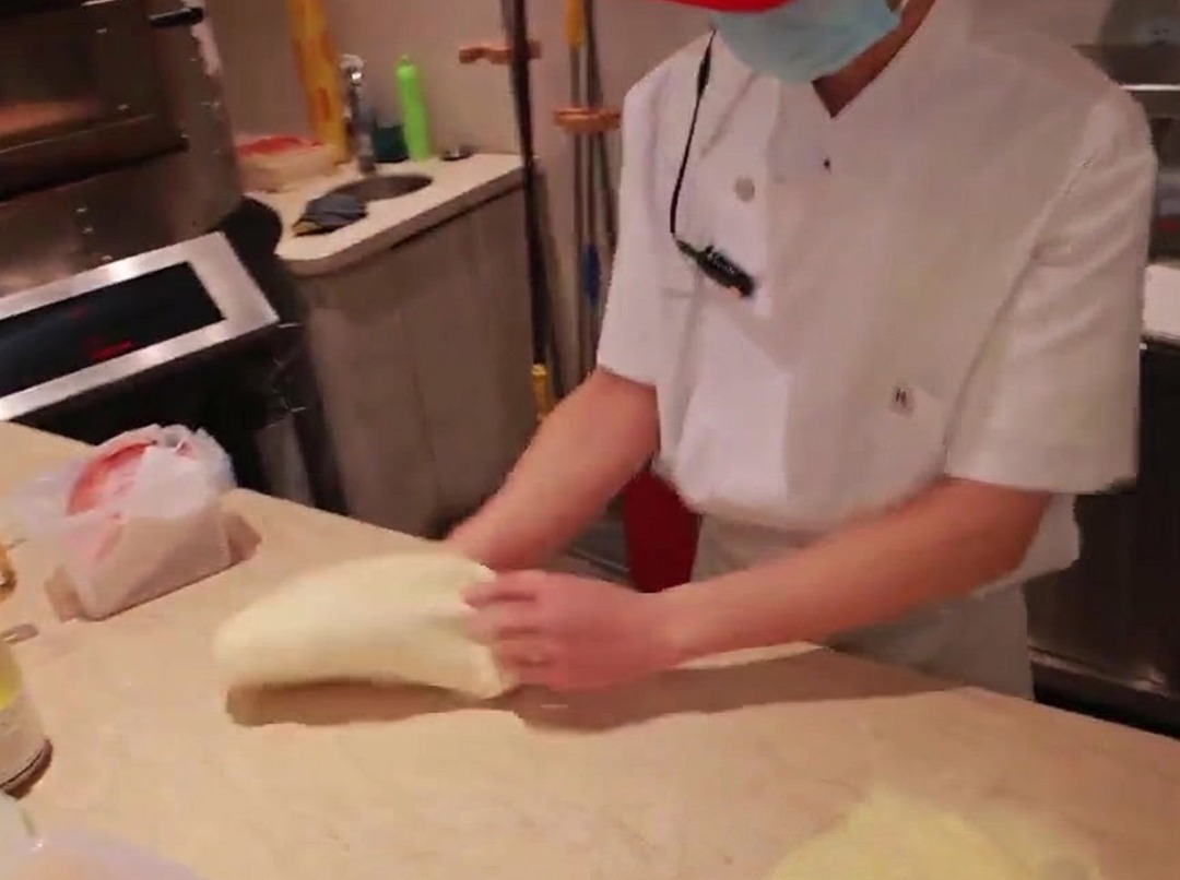 Die Hände eines Kochs beim Ausrollen von Pizzateig auf einer bemehlten Arbeitsfläche.