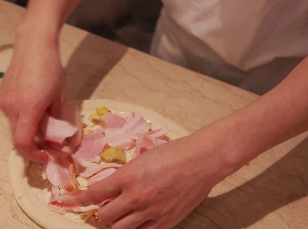 Speckscheiben werden vorsichtig auf die Kartoffelschicht auf dem Pizzateig gelegt.