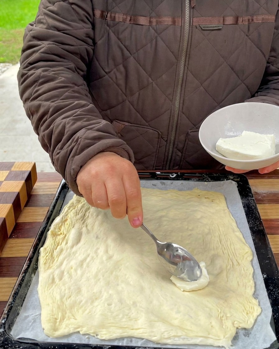 Cremiger Käse wird mit einem Löffel auf dem Pizzateig verteilt.