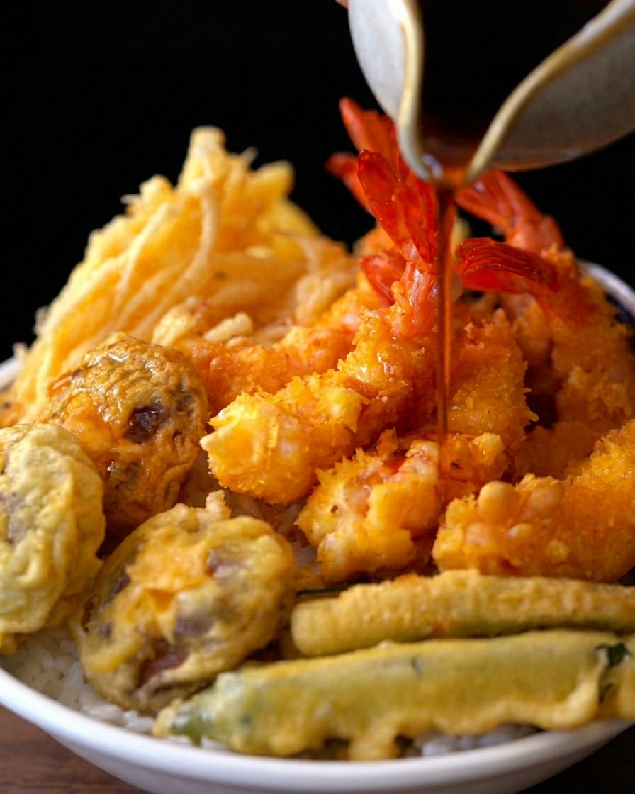 Eine dunkle, herzhafte Sauce wird aus einem Keramikgefäß über eine Schüssel mit Reis, frittierten Garnelen und Gemüse gegossen.
