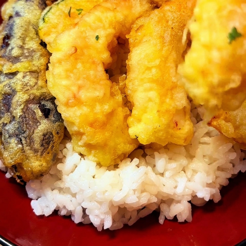 Goldbraun frittierte Tempura-Stücke, einschließlich Garnelen und Gemüse, werden kunstvoll auf einem Bett aus Reis in einer roten Schüssel arrangiert.