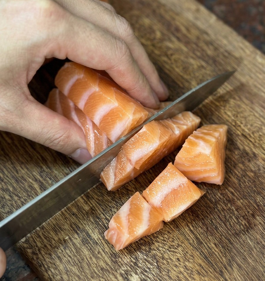 Ein scharfes Messer schneidet frischen, orangefarbenen Lachs in gleichmäßige kleine Würfel auf einem Holzbrett.