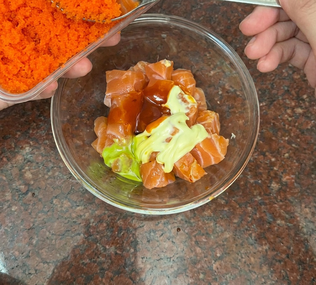 Ein silberner Löffel gibt leuchtend orangefarbenen Fischrogen zu einer Schüssel mit gewürfeltem Lachs.