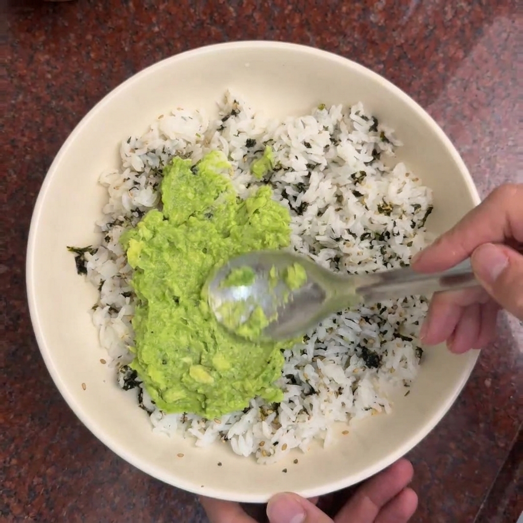 Ein Löffel verteilt hellgrüne, zerdrückte Avocado auf einer Schüssel mit weißem Reis, der mit dunklen Algenstreifen garniert ist.