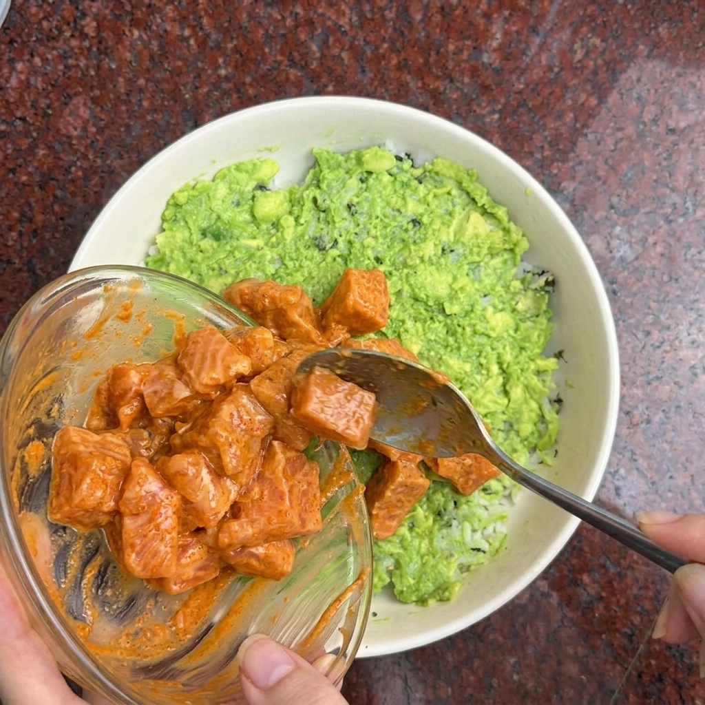 Eine Hand gibt mit einem Löffel in orangefarbener Marinade gewendete Lachswürfel auf eine Reis-Avocado-Bowl.