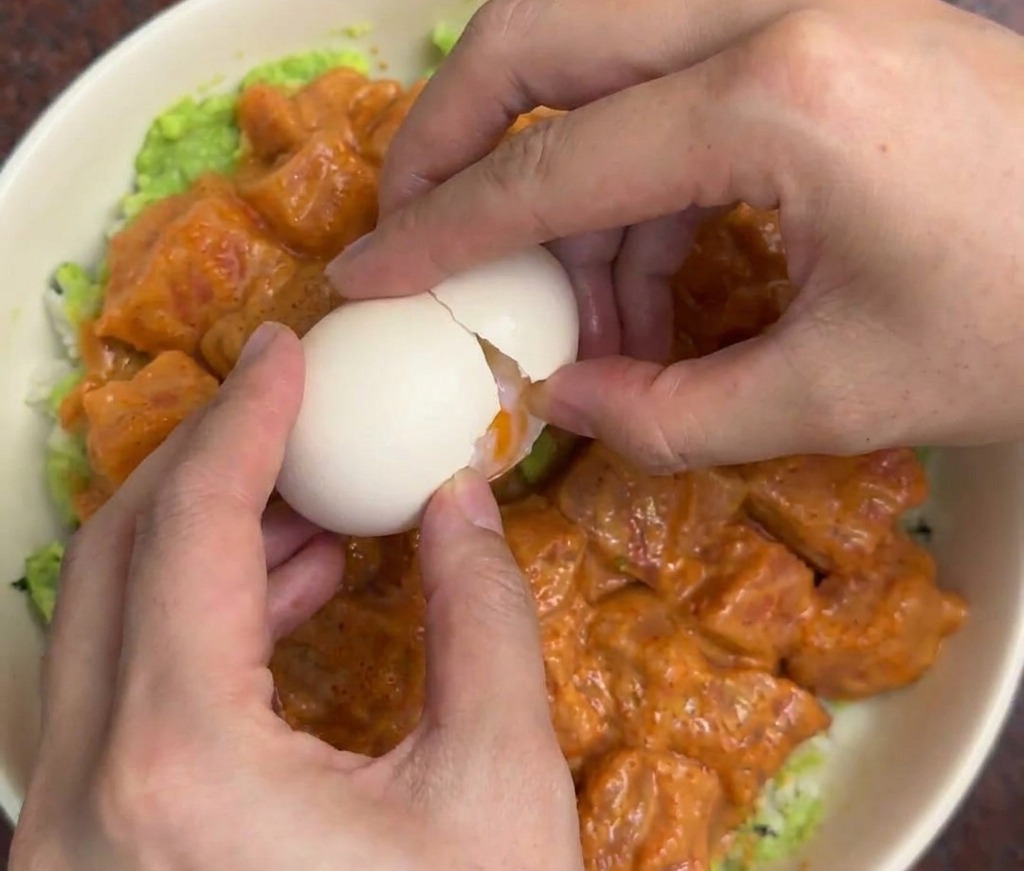 Hände schlagen vorsichtig ein weißes Onsen-Ei in die Mitte einer Lachs-Avocado-Reis-Bowl auf.