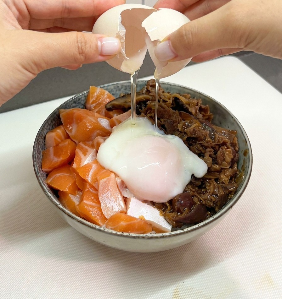 Zwei Hände schlagen vorsichtig eine weiße Eierschale auf, um ein weiches Onsen-Ei auf die Lachs-Rindfleisch-Bowl gleiten zu lassen.