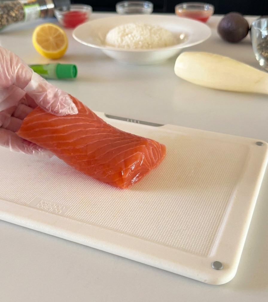 Eine behandschuhte Hand hält ein frisches Lachsfilet auf einem weißen Schneidebrett, bereit, es in Würfel zu schneiden.