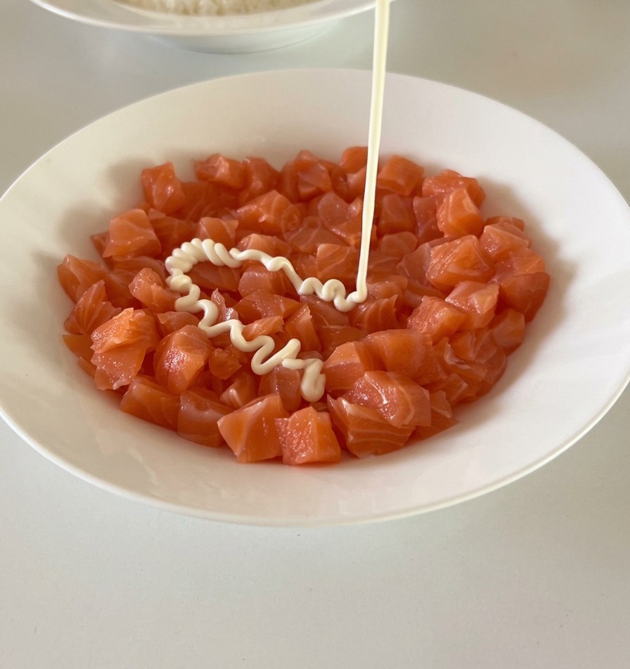 Cremig weiße Mayonnaise wird aus einer Flasche auf eine Schüssel mit frischen, orangefarbenen Lachswürfeln geträufelt.