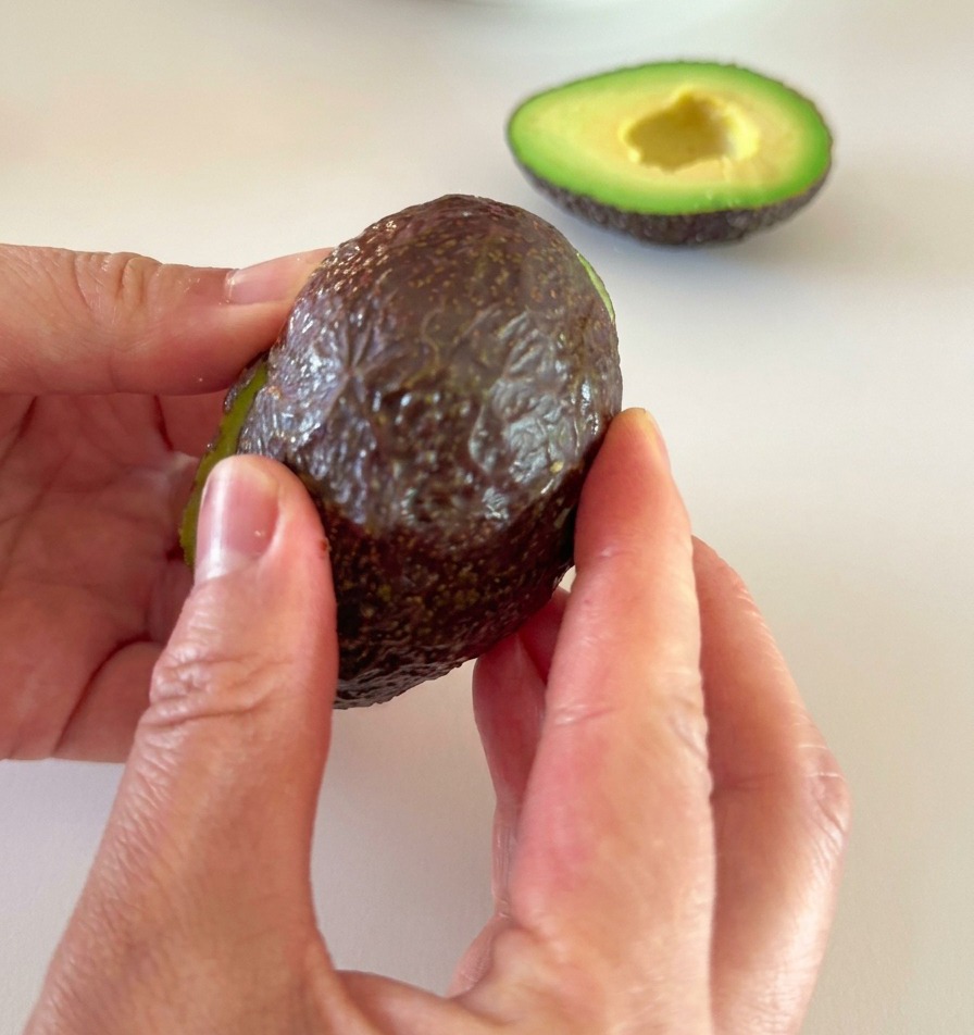 Zwei Hände ziehen vorsichtig die dunkle Schale von einer halbierten, reifen grünen Avocado ab.