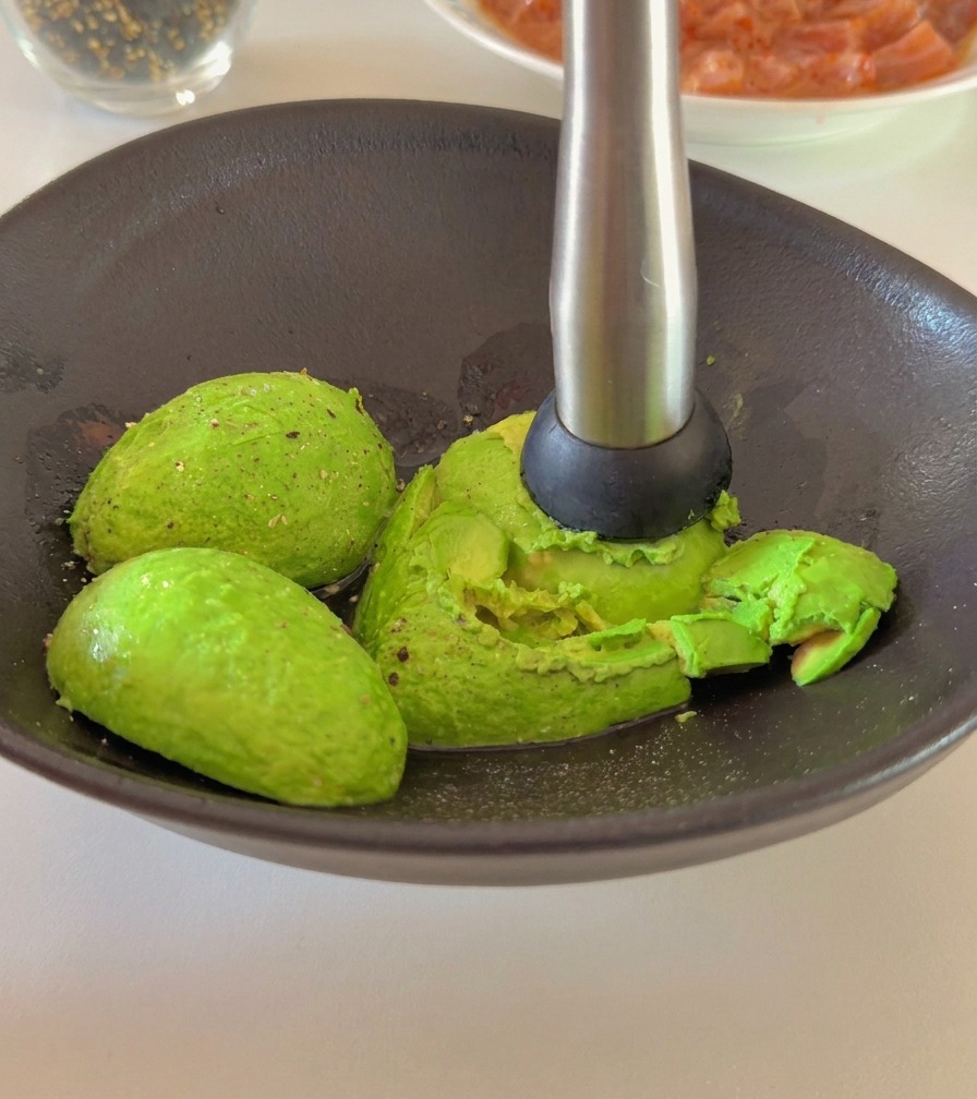 Eine Person zerdrückt mit einem Stampfer aus Metall reife Avocadohälften in einer dunklen Keramikschale.