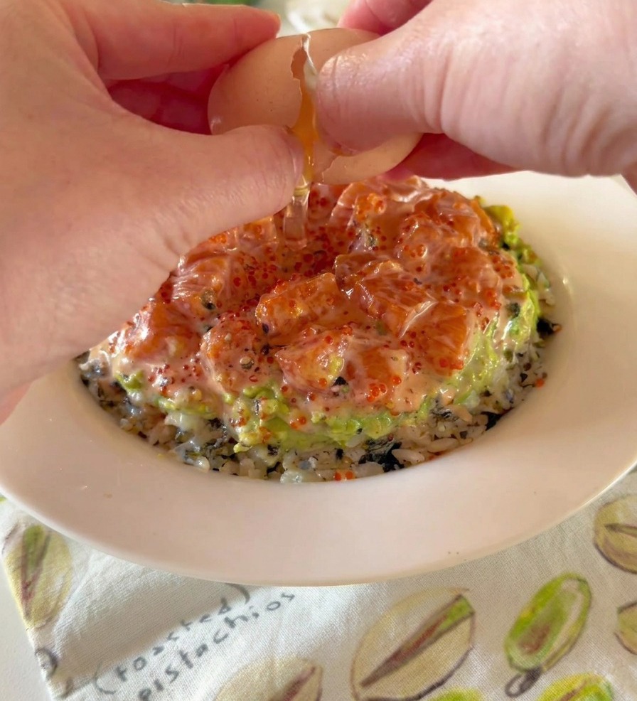 Die Hände einer Person schlagen ein weichgekochtes Onsen-Ei über einer Bowl mit Schichten aus Reis, zerdrückter Avocado und gewürfeltem Lachs auf.