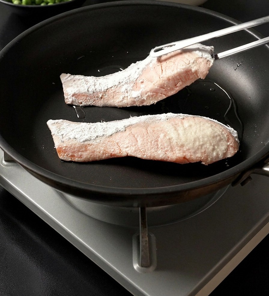 Eine Metallzange legt ein in Stärke gewendetes Lachsfilet neben ein weiteres Filet in eine heiße Pfanne.