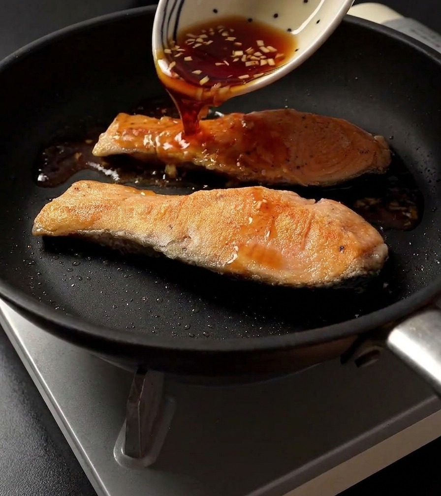 Dunkle Teriyaki-Sauce wird aus einer gemusterten weißen Schüssel auf zwei gebratene Lachsfilets in einer Pfanne gegossen.