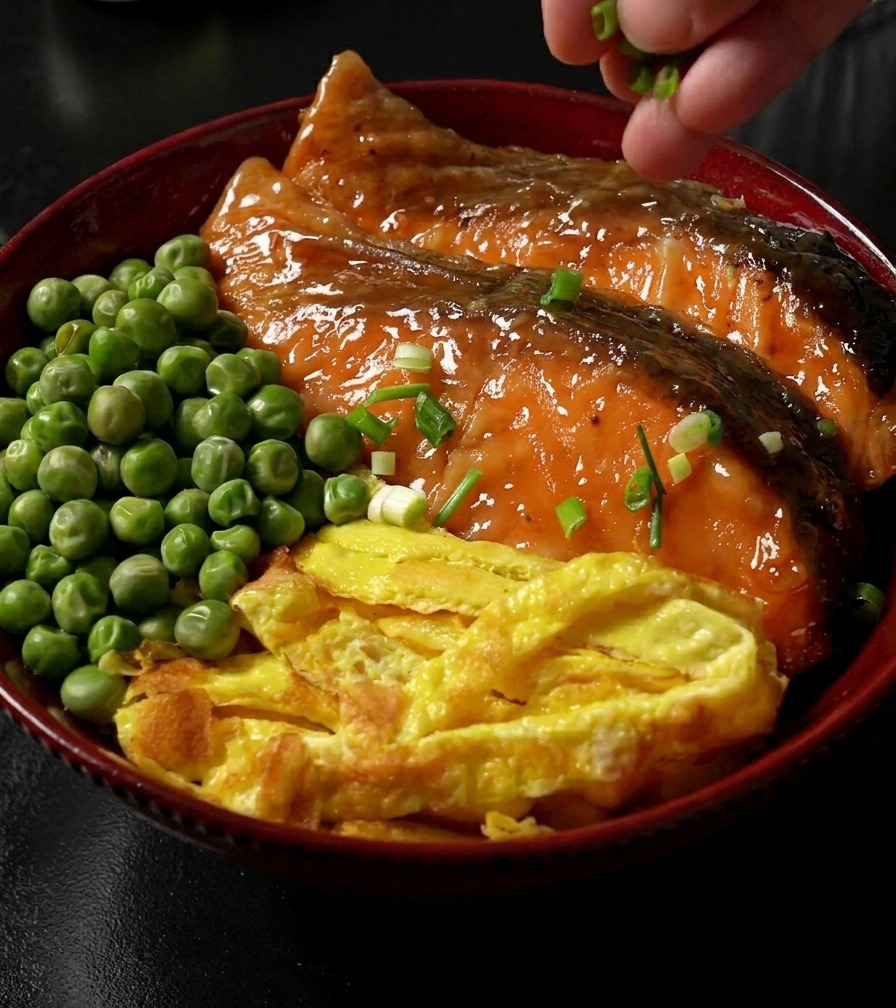 Eine Hand streut gehackte Frühlingszwiebeln über eine fertige Reisschüssel, die mit Teriyaki-Lachs, Eierstreifen und grünen Erbsen garniert ist.