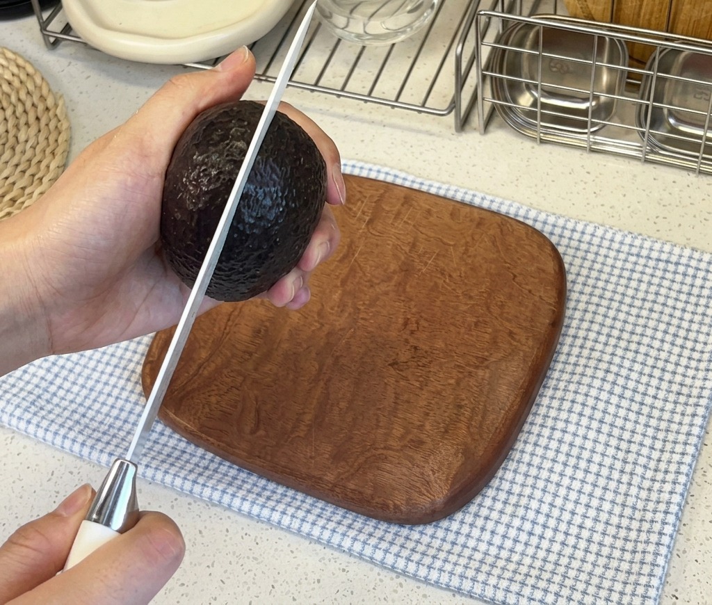 Die Hände einer Person halten eine dunkelhäutige Avocado, während sie diese vertikal mit einem scharfen Küchenmesser aufschneiden.
