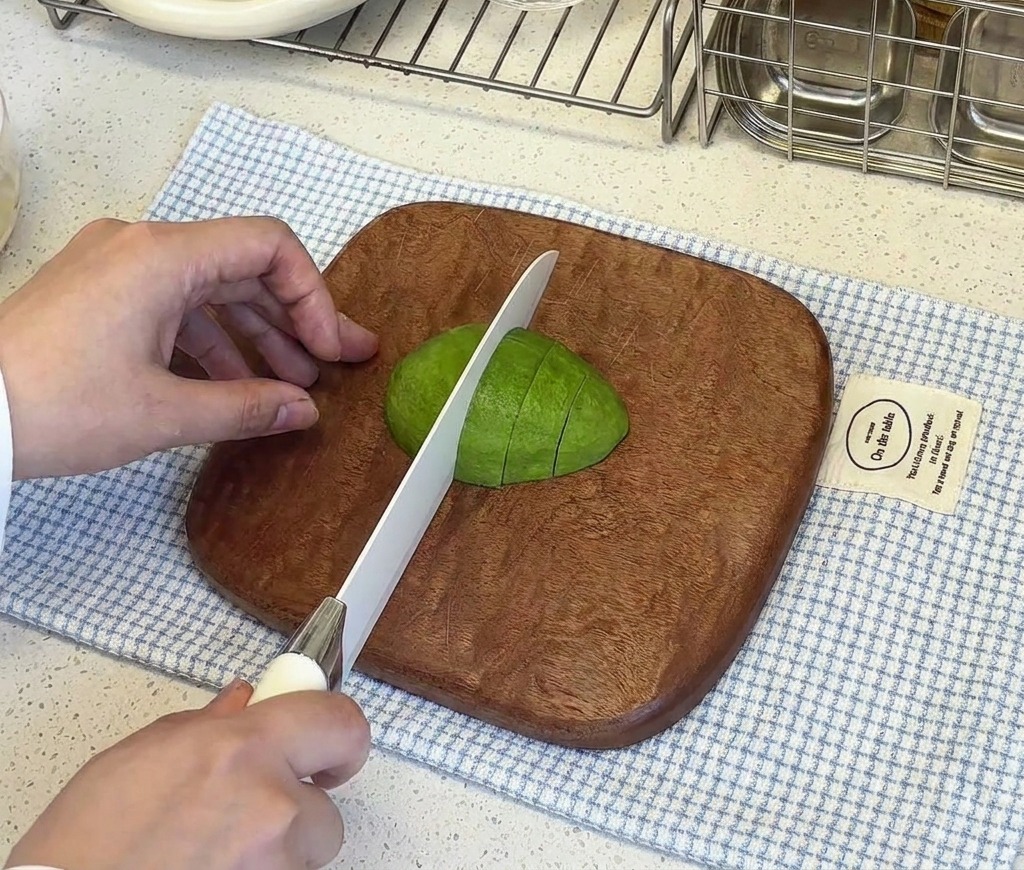 Eine Hand fixiert eine geschälte Avocadohälfte auf einem Holzbrett, während ein weißes Messer sie in kleine Würfel schneidet.