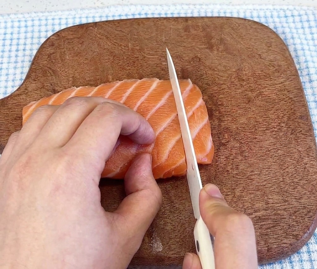 Nahaufnahme eines scharfen Messers, das durch ein frisches, marmoriertes Lachsfilet auf einem dunklen Holzschneidebrett schneidet.