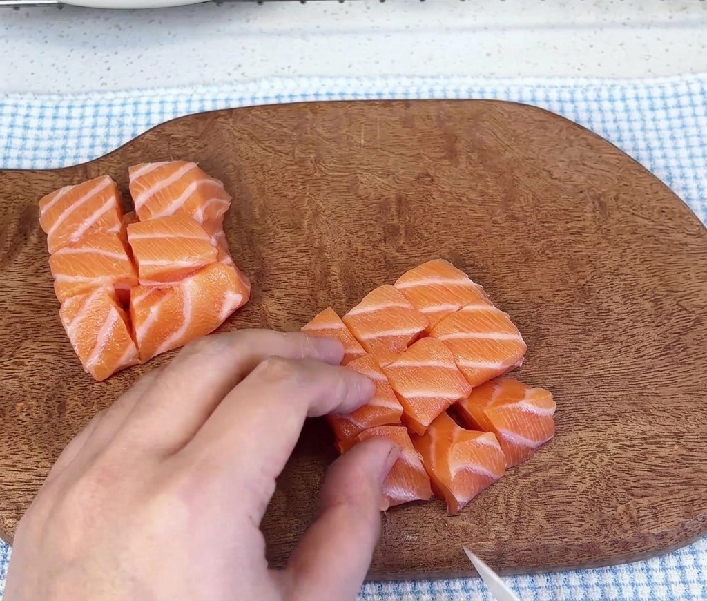 Mehrere Haufen gewürfelter roher Lachswürfel liegen nach dem Schneiden auf einem Holzschneidebrett.