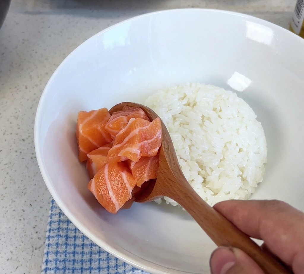 Ein Holzlöffel hebt einen Haufen frischer Lachswürfel an und gibt sie in eine weiße Schüssel mit gedämpftem Reis.