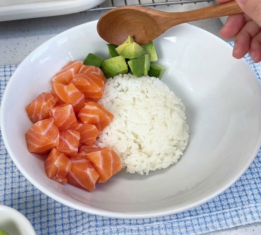 Gewürfelte Avocado wird mit einem Löffel in eine weiße Schüssel neben einen Haufen roher Lachswürfel über Reis gegeben.