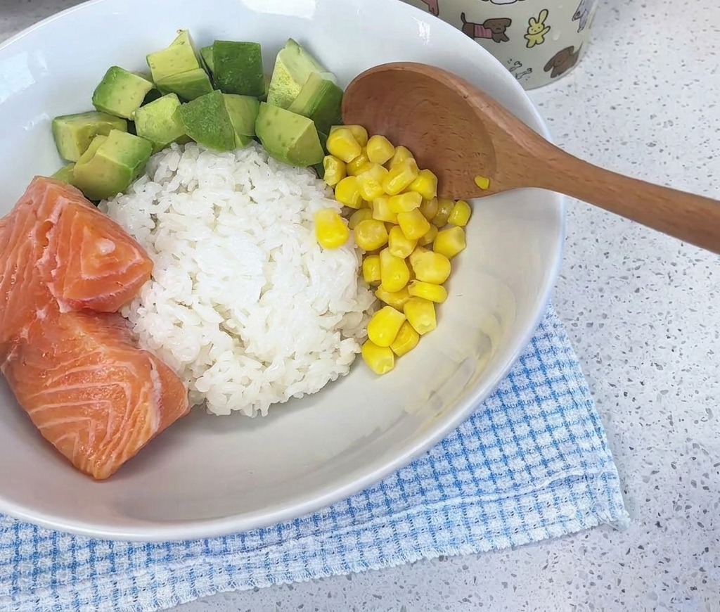 Ein Holzlöffel gibt leuchtend gelbe Maiskörner in eine weiße Schüssel, die mit Reis, gewürfelter Avocado und frischen Lachsstücken gefüllt ist.