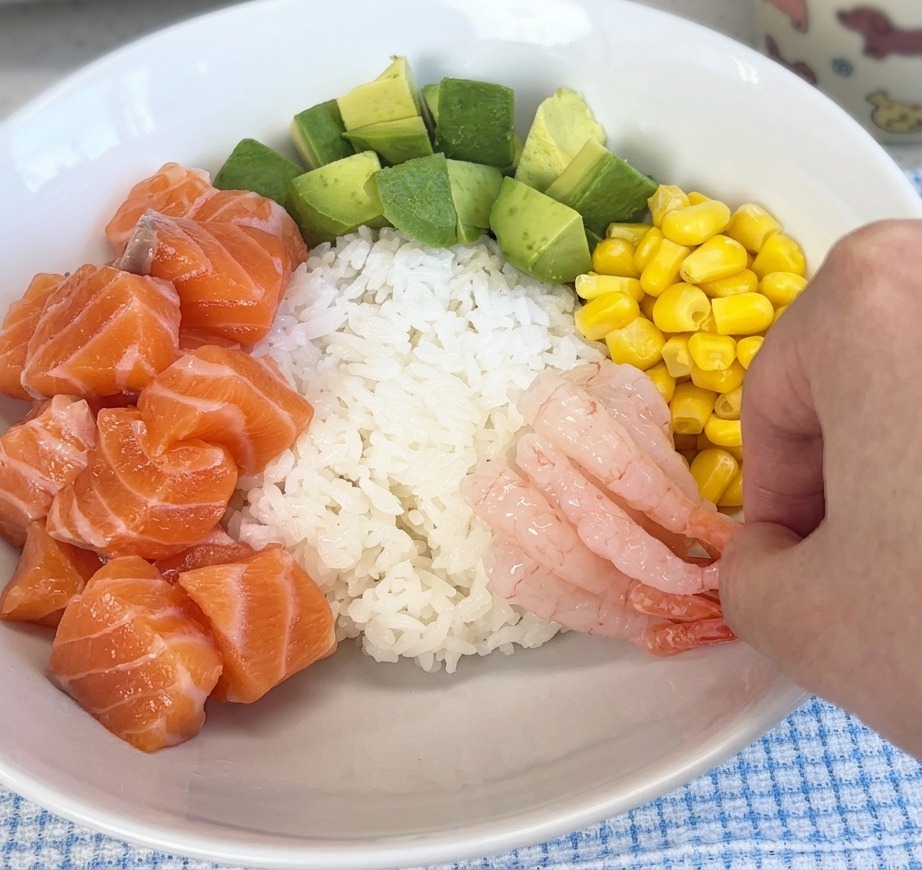 Eine Hand legt rosa, glasige süße Garnelen auf den Reis in einer Schüssel neben Lachs und Mais.
