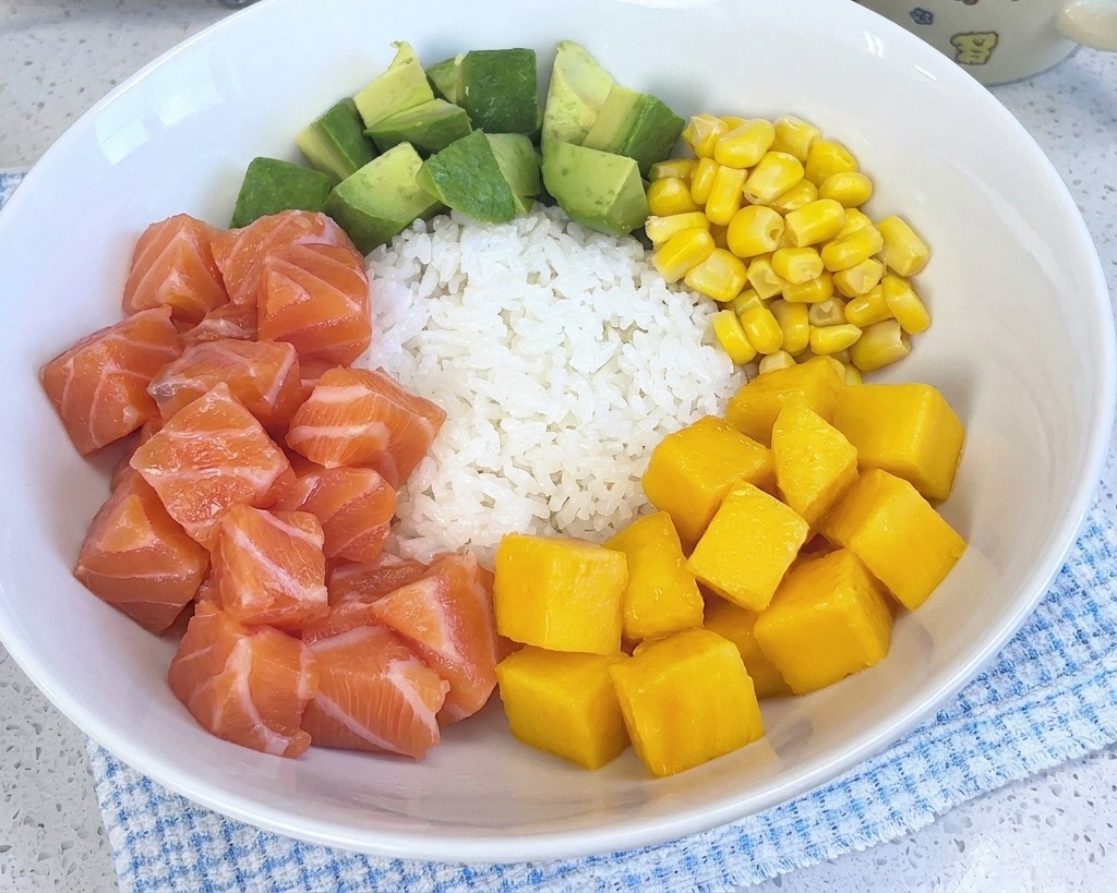 Eine farbenfrohe Poke-Bowl mit weißem Reis, Lachs, Avocado, Mais und Garnelen, die jetzt einen Haufen leuchtend gelber Mangowürfel enthält.