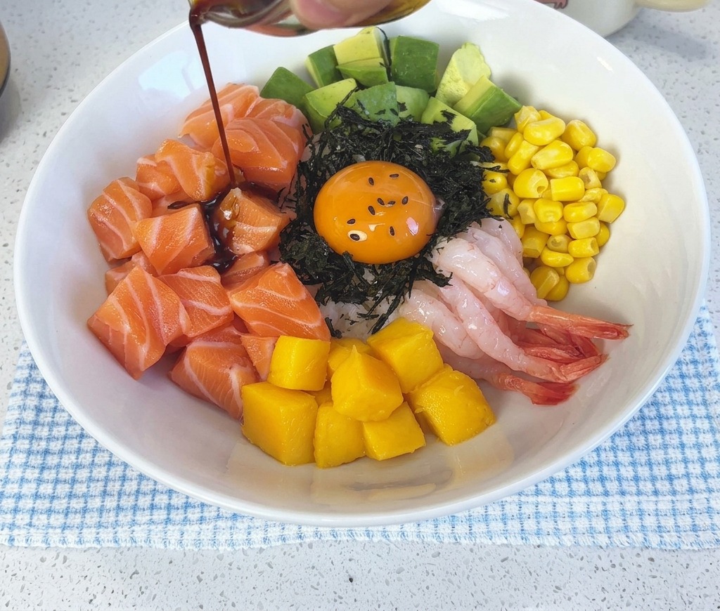 Dunkle Wasabi-Sojasauce wird aus einer kleinen Glasschale über die fertig angerichtete Lachs-Poke-Bowl und das Eigelb gegossen.