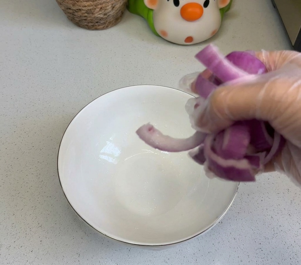 Eine Person gibt geschnittene rote Zwiebeln in eine weiße Keramikschüssel auf einer sauberen Küchenarbeitsplatte.