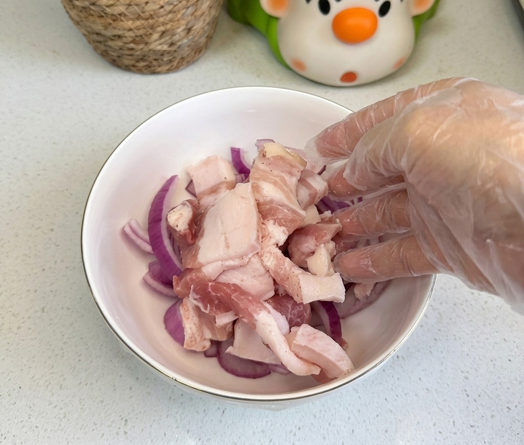 Rohe Schweinebauchscheiben werden in eine weiße Schüssel mit dünn geschnittenen roten Zwiebeln gegeben.