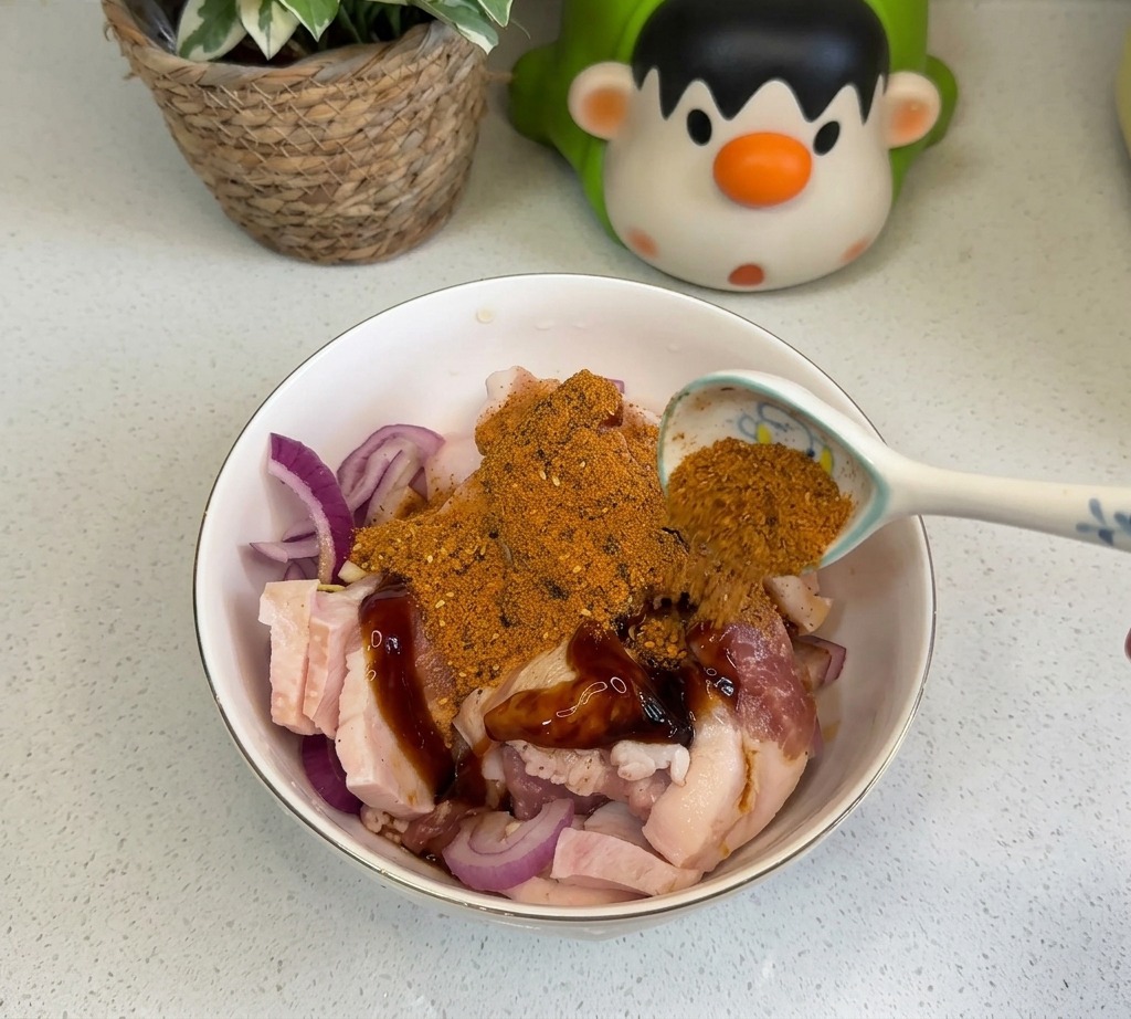 Ein Löffel orangefarbenes BBQ-Gewürz und rotes Chilipulver werden über den marinierten Schweinebauch und die Zwiebeln gestreut.